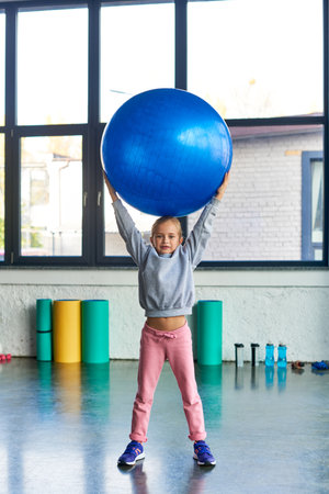 little cute girl in sportswear holding fitness ball above head and looking at camera, child sportの写真素材