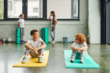 two cheerful boys smiling at each other on fitness mats with cute girls standing on backdropの写真素材