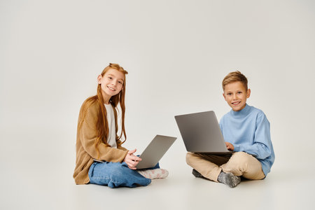 jolly little children in stylish attires on floor with laptops smiling at camera, fashion concept