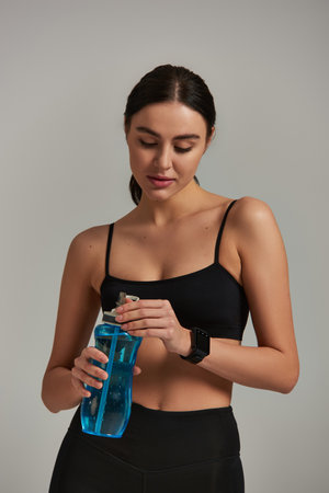 fit woman in leggings and crop top holding bottle with water while standing on grey background