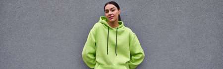 beautiful sportswoman in trendy lime color hoodie standing near grey concrete wall outdoors, banner