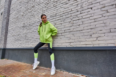 stylish young sportswoman in lime color hoodie, leggings and sneakers standing near grey wall