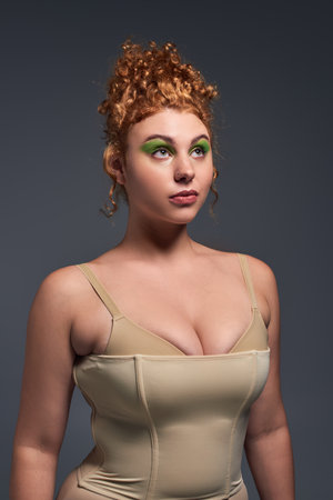 dreamy plus size woman with bold makeup and red hair wearing beige lingerie and looking away on greyの写真素材
