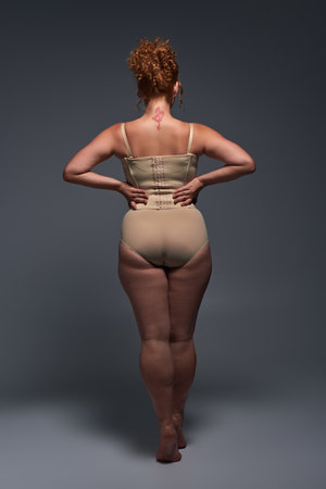 back view of curvaceous redhead woman in beige lingerie with hands on waist on dark grey backdropの写真素材