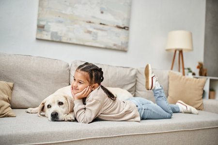 happy girl smiling and lying on sofa with cute labrador in modern living room, pet and childの写真素材