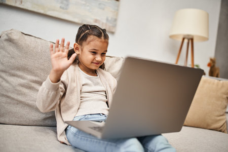 happy elementary age girl sitting on sofa and waving hand at laptop in living room, online lessonの写真素材