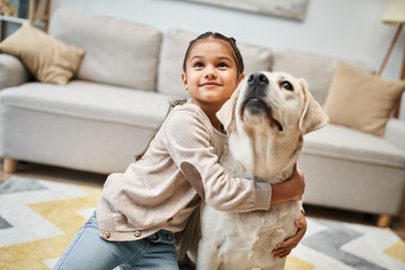 happy cute elementary age girl looking up and hugging labrador in modern apartment, pet and child