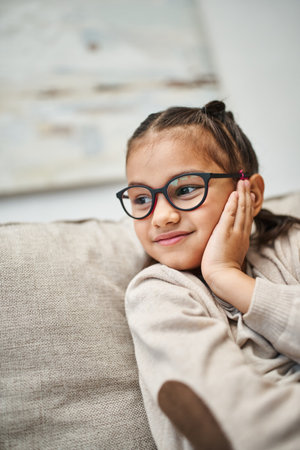 happy elementary age girl in casual attire wearing eyeglasses and looking away in living roomの写真素材