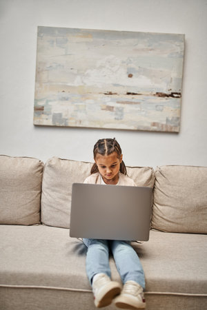 happy elementary age girl sitting on sofa and using laptop in modern living room, e-learning