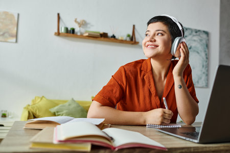 cheerful young student in orange shirt listening music while studying at laptop, looking away