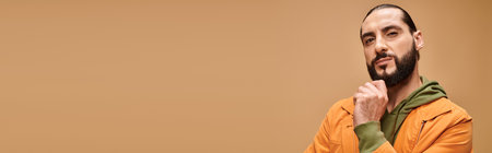 pensive middle eastern man touching beard and standing in casual attire on beige background, banner