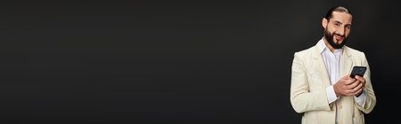 cheerful and bearded arabic man in white shirt and blazer texting smartphone on black, bannerの写真素材