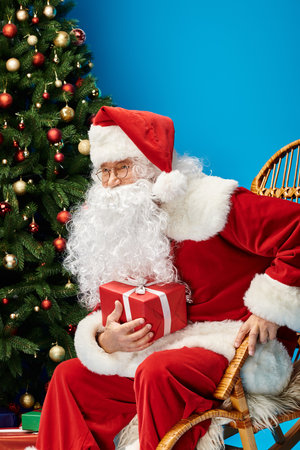 happy Santa Claus with beard and eyeglasses sitting in rocking chair with gift near Christmas treeの写真素材