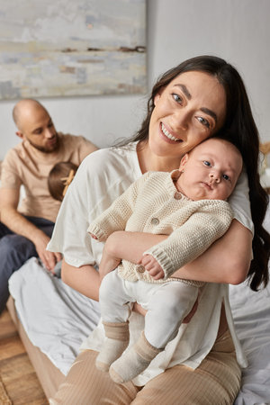 focus on mother holding newborn baby and smiling at camera with blurred husband and son on backdrop