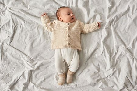 top view of adorable newborn baby boy in beige warm cardigan lying on white blanket with raised arms