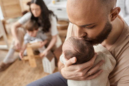 focus on bearded father kissing his baby in forehead with his blurred wife and son on backdropの写真素材