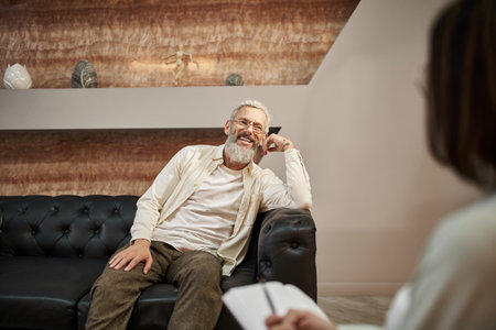 cheerful bearded middled aged man with tattoo sitting on leather couch and looking at psychologist