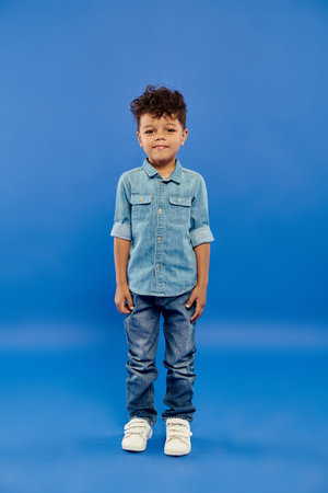 cute and curly african american preschooler boy in stylish denim outfit posing on blue backgroundの写真素材