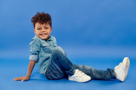 positive african american preschooler boy in stylish denim attire sitting on blue background