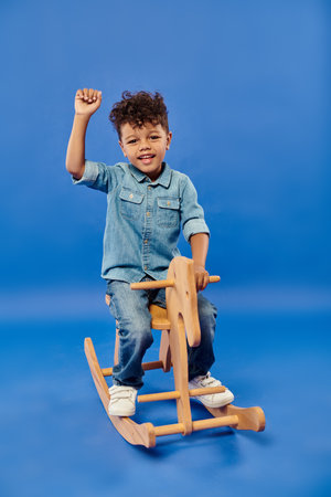 cute african american preschooler boy in stylish denim clothes sitting on rocking horse on blueの写真素材