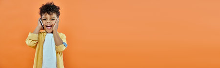 happy african american preschooler boy in casual attire talking on smartphone on orange, banner