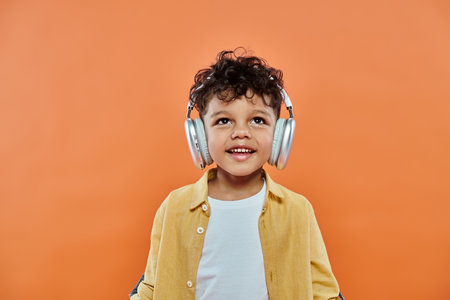 happy african american boy listening music in wireless headphones on orange background, look upの写真素材