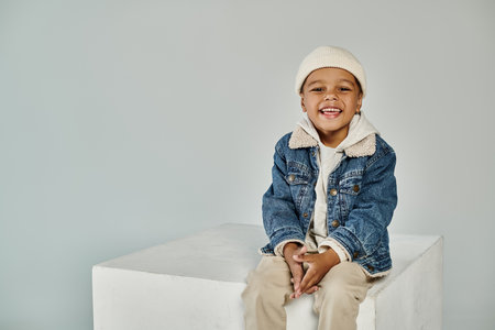 cute joyful african american boy in winter attire and beanie hat sitting on concrete cube on grey