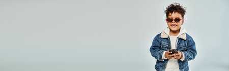 happy african american boy in winter attire and sunglasses using smartphone on grey backdrop, bannerの写真素材