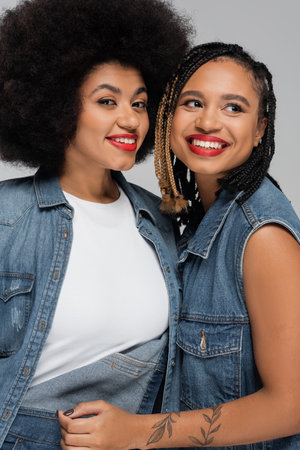 tattooed and joyful african american woman in denim clothes smiling near trendy girlfriend on grey