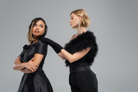 elegant woman in black touching shoulder of african american girlfriend with folded arms on greyの写真素材