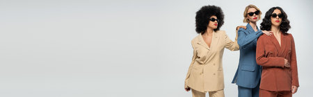 multiracial girlfriends in suits and sunglasses touching shoulders of each other on grey, banner