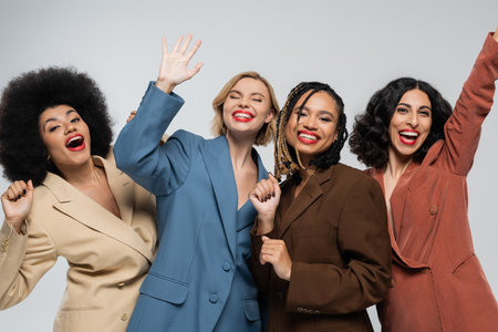 overjoyed multiracial girlfriends in colorful suits waving hands on grey, fashion and happinessの写真素材