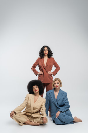 stylish multiracial woman with hands on hips near girlfriends in colorful suits sitting on greyの写真素材