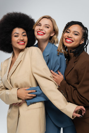 happy multiracial girlfriends in multicolored suits embracing on grey, togetherness and styleの写真素材