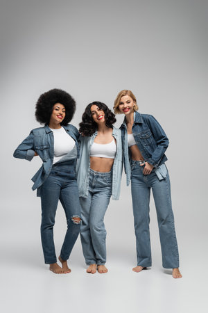 full length of barefoot multiracial girlfriends in blue denim clothes posing on grey backdropの写真素材