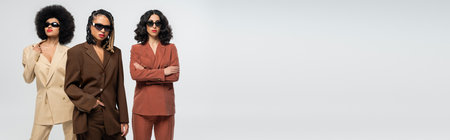 confident multiracial girlfriends in dark sunglasses and trendy colorful suits on grey, bannerの写真素材