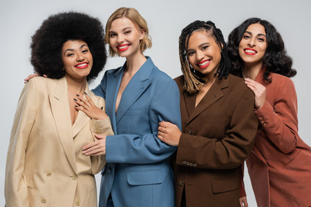 four jolly multiracial girlfriends in trendy colorful suits smiling at camera on grey, diversityの写真素材