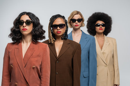 diverse group of fashionable multiracial girlfriends in dark sunglasses and colorful suits on greyの写真素材