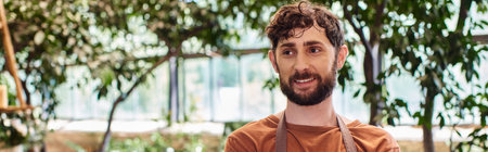professional headshots, cheerful bearded gardener in apron and gloves posing in greenhouse, bannerの写真素材