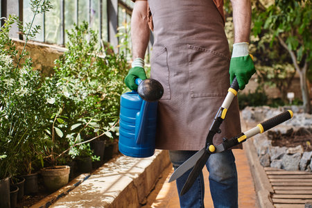 cropped gardener in gloves and apron holding big gardening scissors and watering can in greenhouse