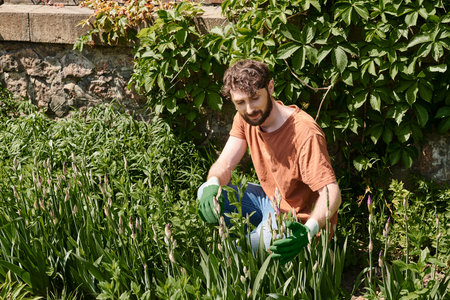 pleased and bearded gardener in sun hat examining plant near modern greenhouse in countrysideの写真素材
