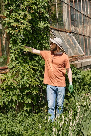 handsome and bearded gardener in sun hat examining plant near modern greenhouse in countrysideの写真素材