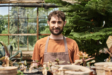 happy bearded gardener in linen apron looking at camera around plants in greenhouse, horticultureの写真素材