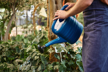 good looking and bearded gardener in denim apron watering plants in greenhouse, horticulture conceptの写真素材