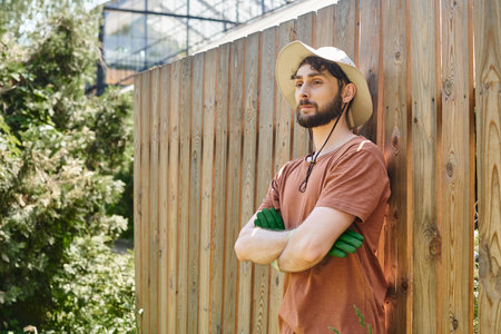 handsome and bearded farmer in sun hat standing with crossed arms near fence in countrysideの写真素材