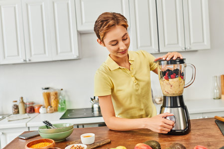 smiling woman grinding fresh fruits in blender while making delicious vegetarian smoothie in kitchen