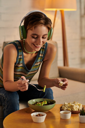joyful woman in headphones having dinner with vegetarian salad and tofu cheese on sofa at home