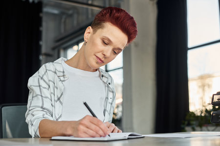 concentrated stylish bigender person sitting at workplace in modern office and writing in notebookの写真素材