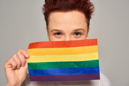 joyful redhead queer person obscuring face with small LGBT flag and looking at camera on greyの写真素材