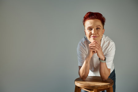 positive bigender person in white t-shirt posing with stool in sunlight looking at camera on greyの写真素材